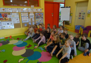 Dzieci siedzą skrzyżnie w grupie na dywanie. Twarze mają zwrócone w kierunku tablicy interaktywnej.